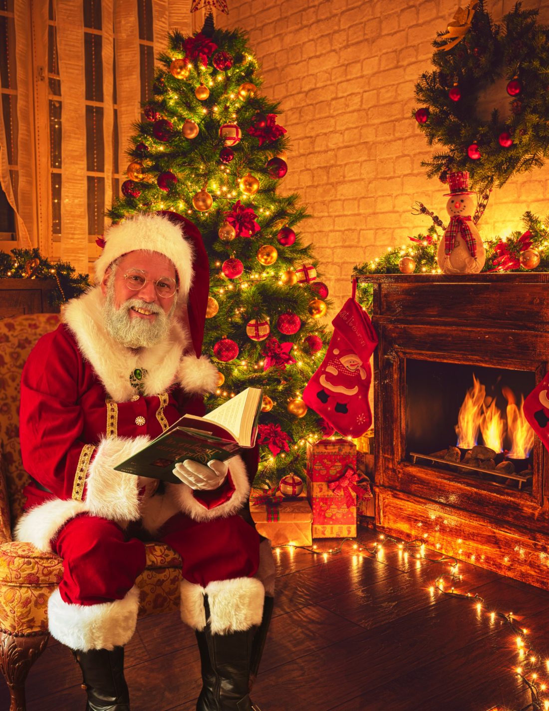 Santa seated by a lit Christmas tree holding a storybook